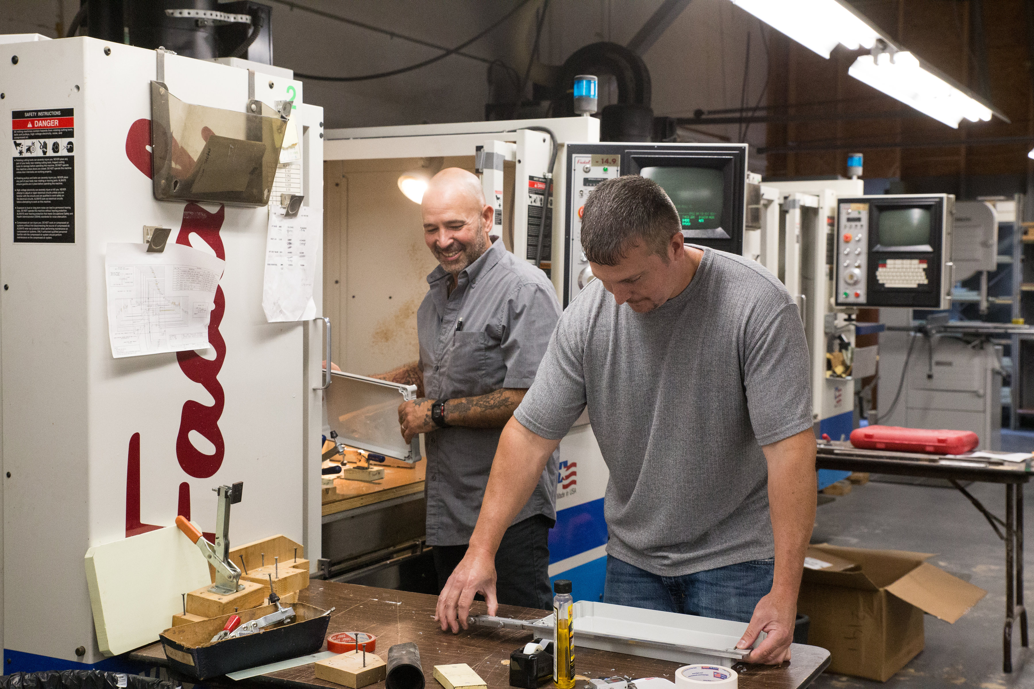 Two AMI employees working at CNC machines in a manufacturing shop, one measuring a metal part on a workbench
