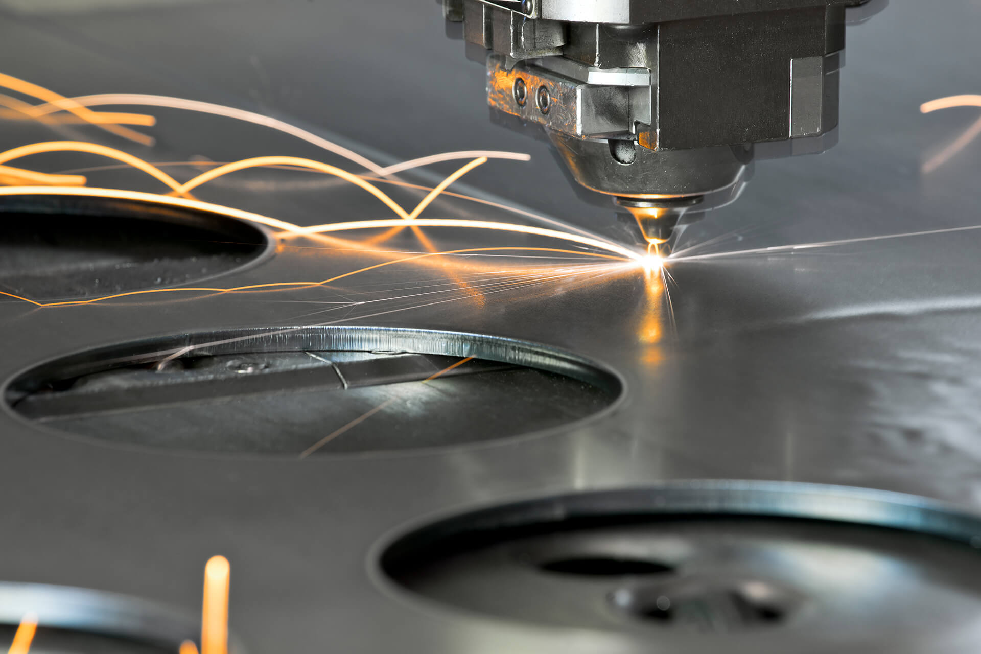 Close-up of laser cutting head slicing metal sheet, producing bright sparks and smooth circular cutouts