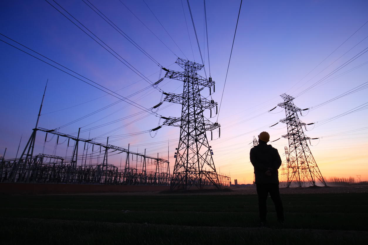 Silhouetted electricity worker near high-voltage pylons and power lines at sunset, with substation structures in the distance