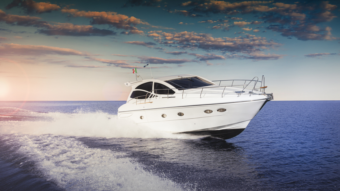 White luxury motor yacht speeding across calm blue sea at sunset, leaving a foamy wake under a dramatic clouded sky