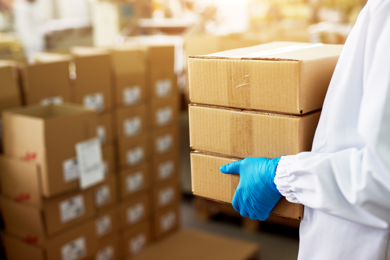Worker in blue gloves and white coat carrying a stack of taped brown boxes in a warehouse with cartons stacked in background