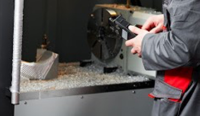 Machinist measuring a metal part beside a CNC lathe chuck, with metal shavings covering the machine bed