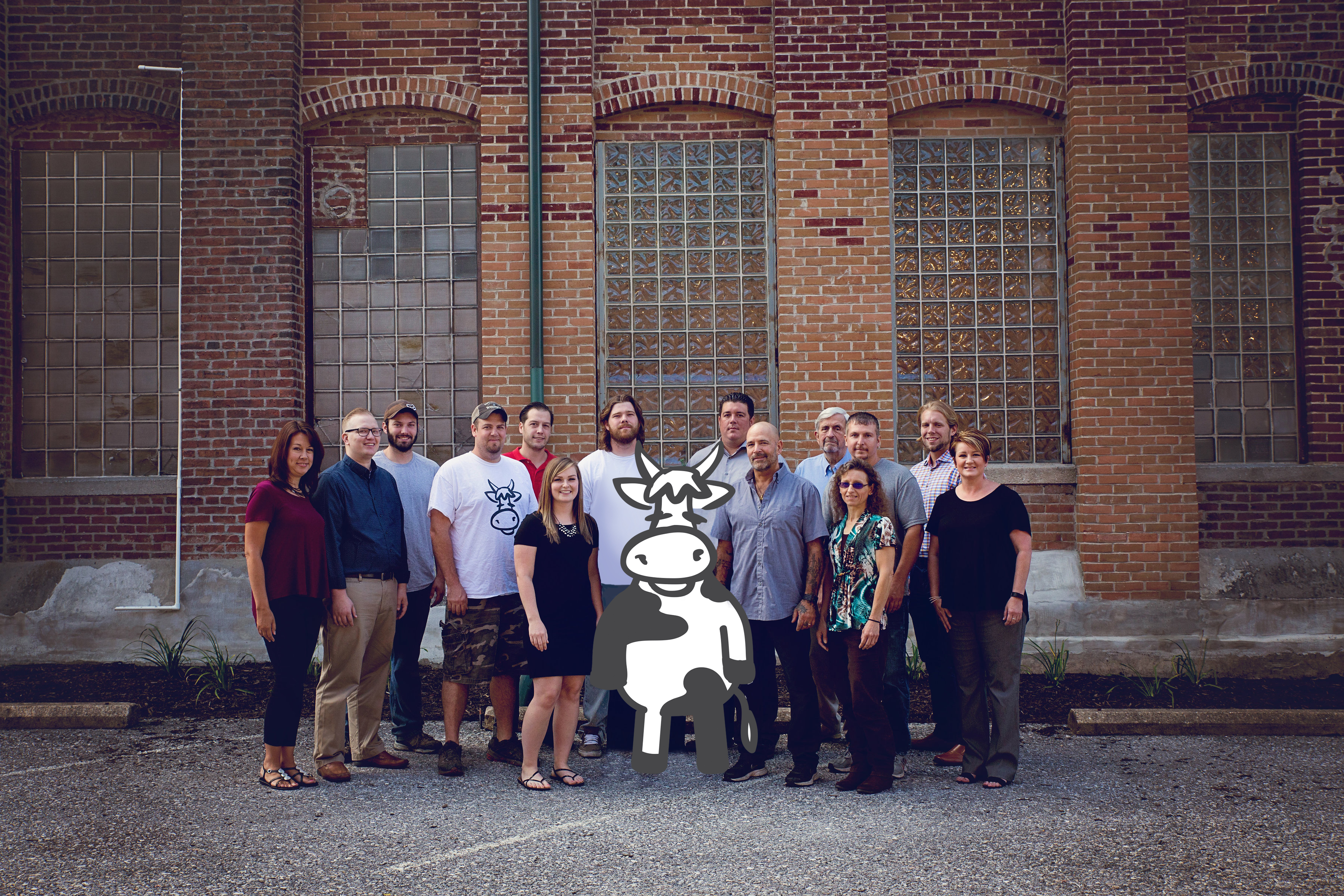Group of coworkers posing outdoors in front of a brick building with glass-block windows, with a large cow graphic centered in front