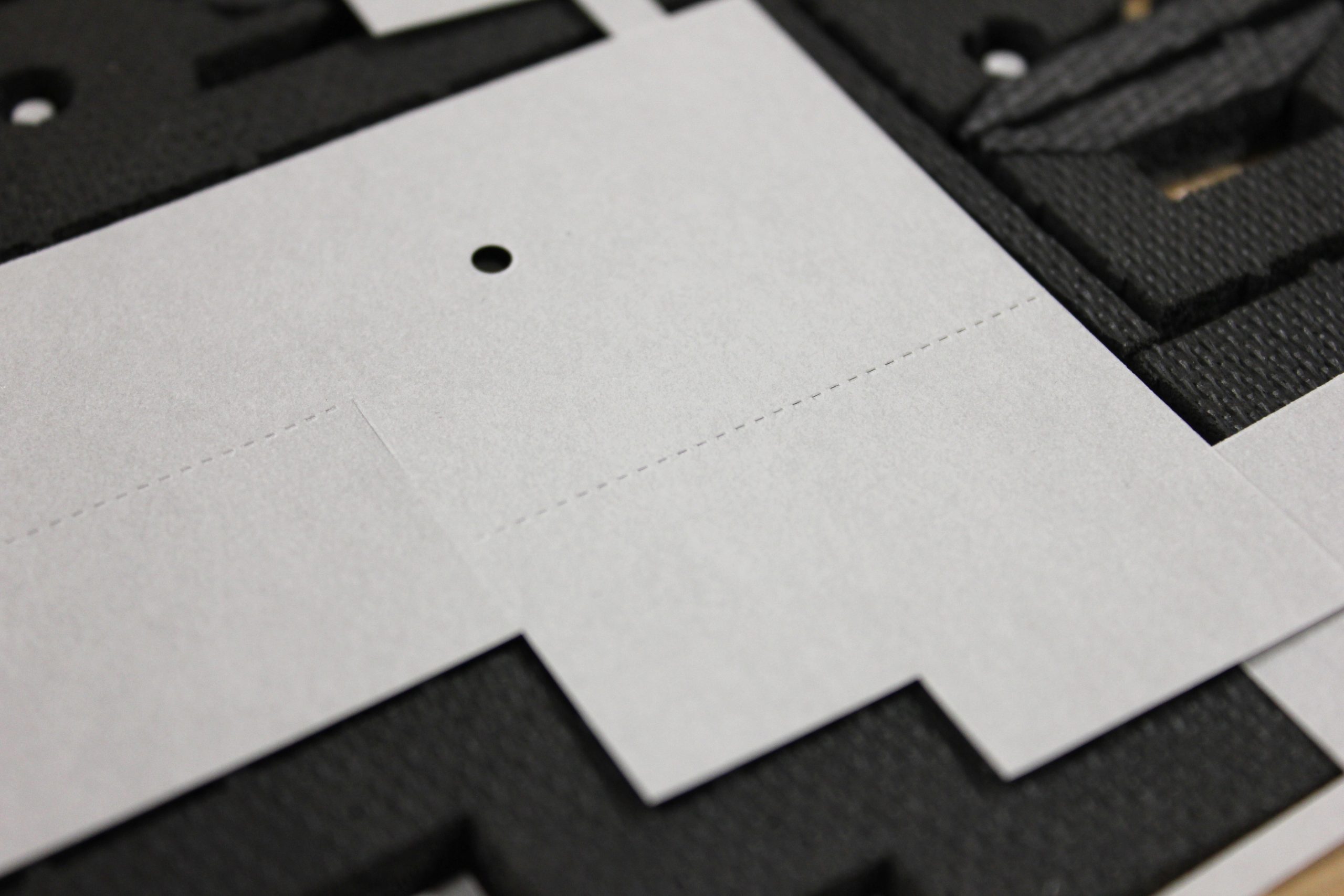 Close-up of white paperboard with a round punched hole and dotted perforation lines on a dark textured surface