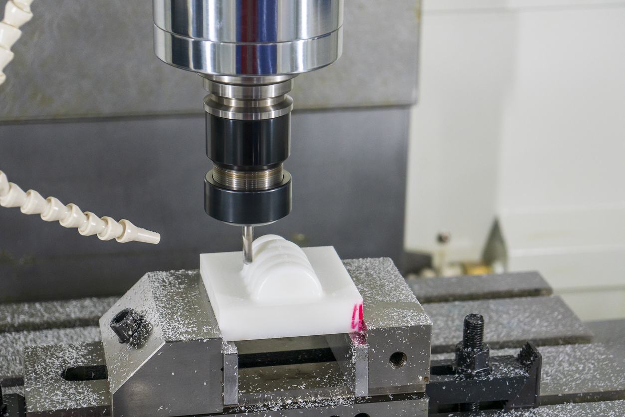 CNC milling machine spindle cutting a white plastic workpiece clamped in a vise, with coolant hose and machining chips visible