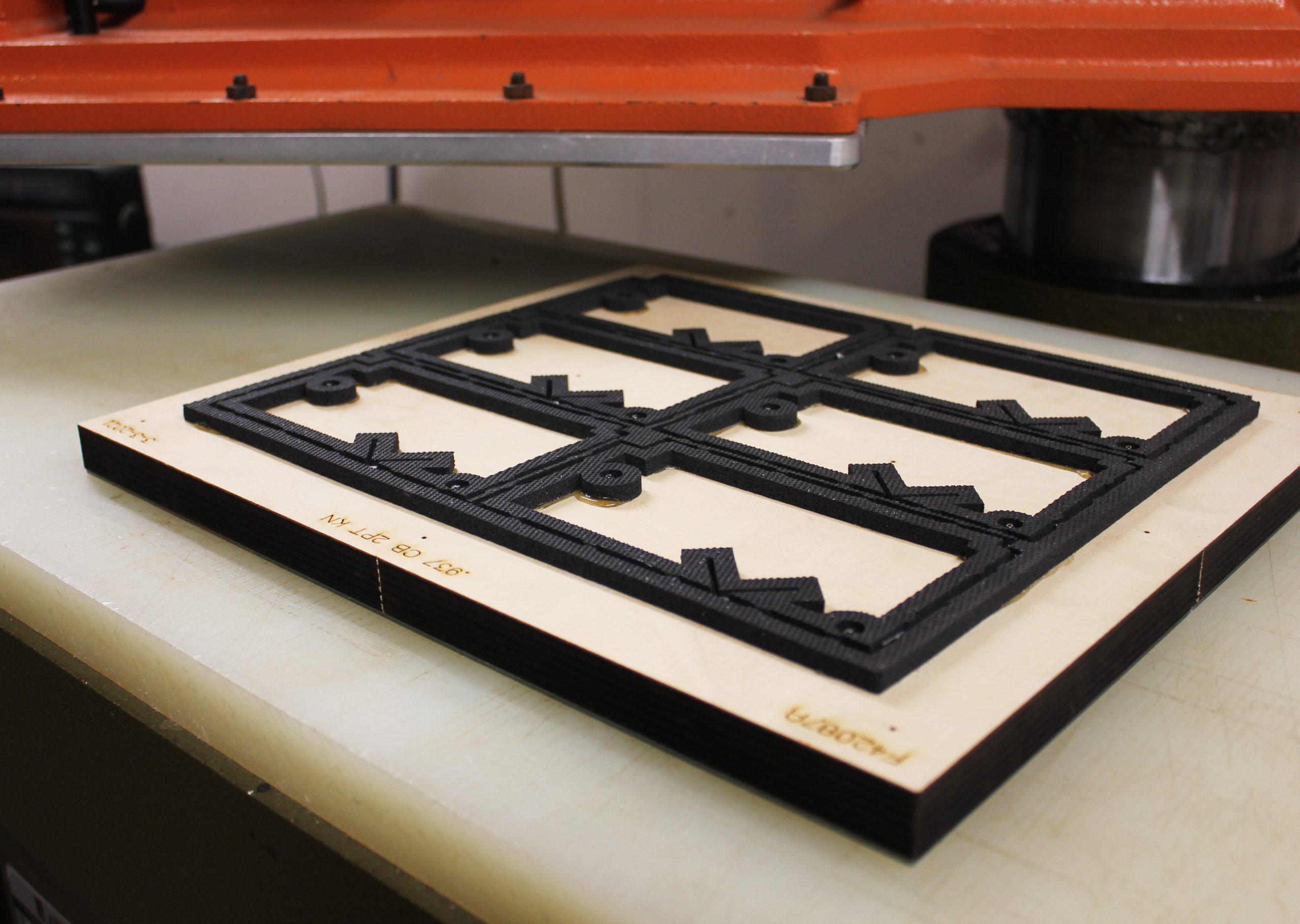 Steel rule die cutting form on a wooden board beneath a press, showing black die layout with multiple rectangular cutouts