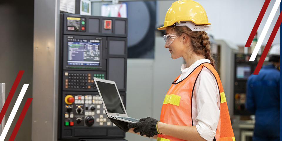 Factory worker in hard hat using laptop beside CNC machine control panel for monitoring and data logging